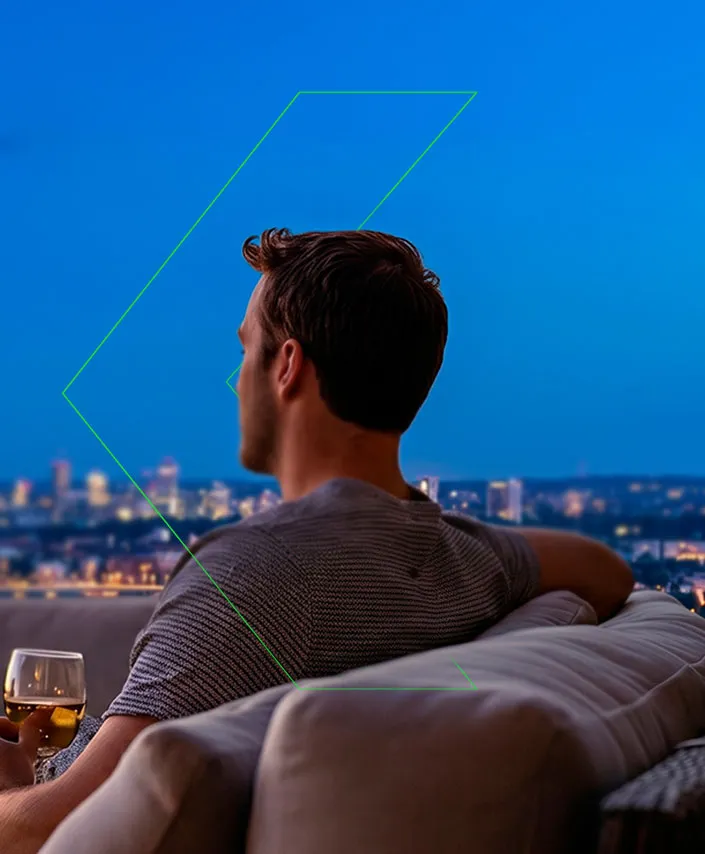 Hombre con copa de vino relajado viendo la vista nocturna de la ciudad desde un sofá al aire libre.
