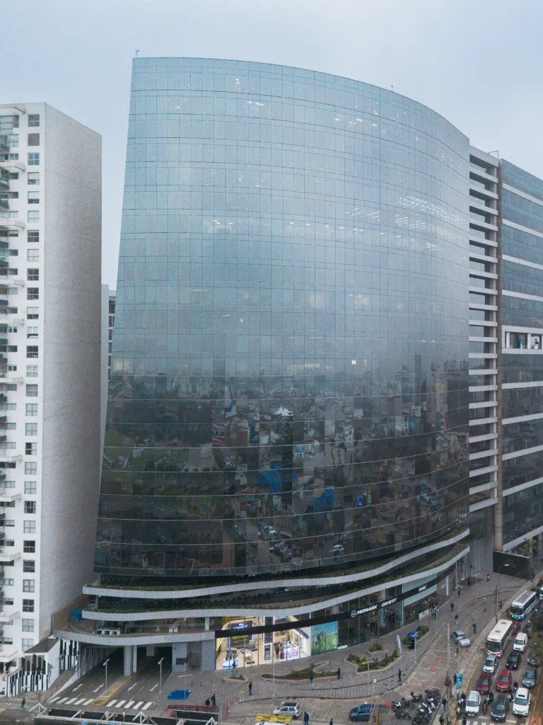 Edificio moderno de vidrio en área urbana, reflejando el cielo nublado y edificios cercanos en sus ventanas.