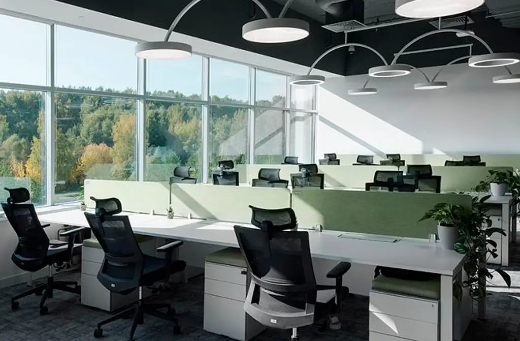 Oficina moderna con escritorio compartido, sillas ergonómicas y grandes ventanales con vista al exterior verde.