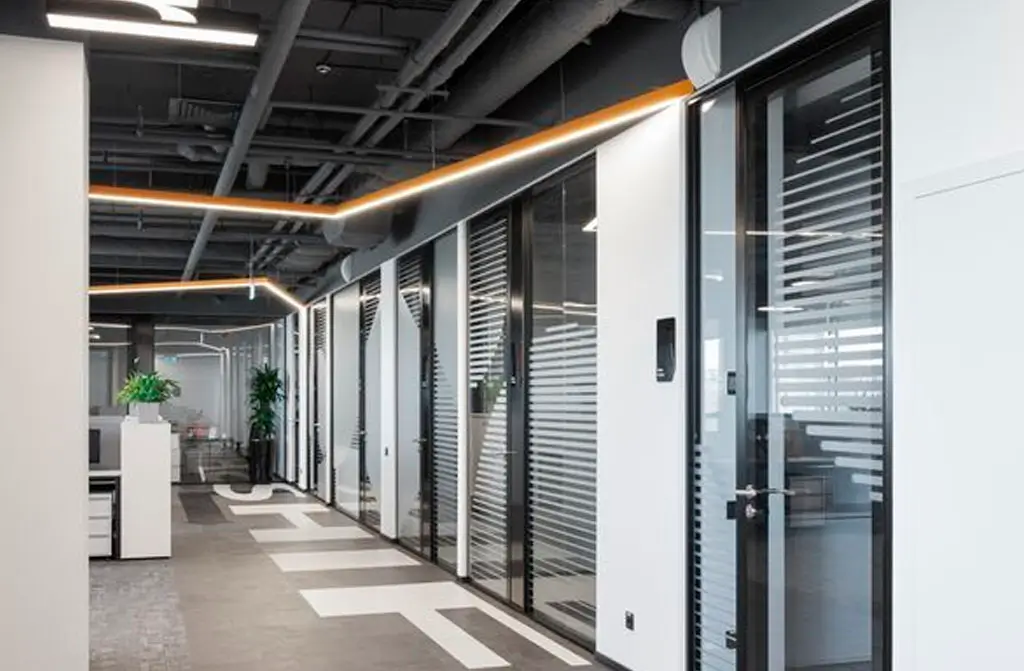 Oficina moderna con paredes de cristal y plantas decorativas, iluminada con luces LED en el techo industrial.